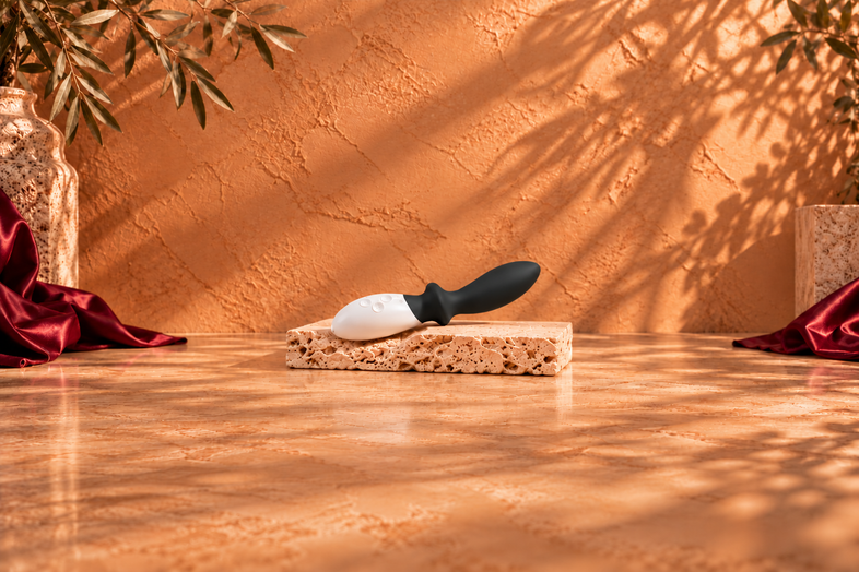 Black and white pleasure massager resting on a light travertine slab, with a warm terracotta background, botanical shadows and burgundy satin