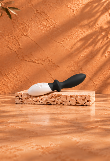Black and white pleasure massager resting on a light travertine slab, with a warm terracotta background, botanical shadows and burgundy satin