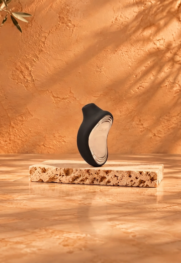 Black LELO SONA 2 on light travertine against a warm copper-beige background with burgundy satin accents