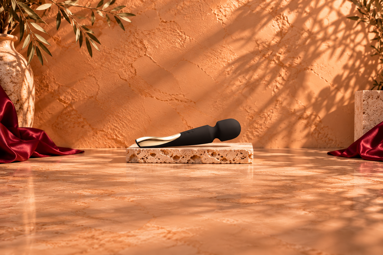 Black wand massager with a gold-coloured detail resting on a light travertine slab, against a warm terracotta background with botanical shadows and burgundy satin