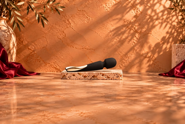 Black wand massager with a gold-coloured detail resting on a light travertine slab, against a warm terracotta background with botanical shadows and burgundy satin