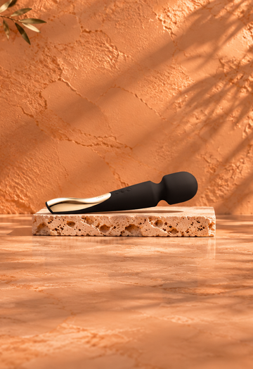Black wand massager with a gold-coloured detail resting on a light travertine slab, against a warm terracotta background with botanical shadows and burgundy satin