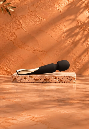 Black wand massager with a gold-coloured detail resting on a light travertine slab, against a warm terracotta background with botanical shadows and burgundy satin