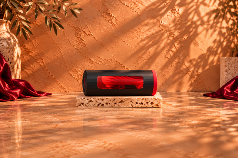 Black and red LELO masturbator resting horizontally on a light travertine slab, against a warm terracotta background with botanical shadows and burgundy satin