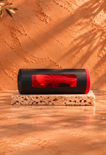 Black and red LELO masturbator resting horizontally on a light travertine slab, against a warm terracotta background with botanical shadows and burgundy satin