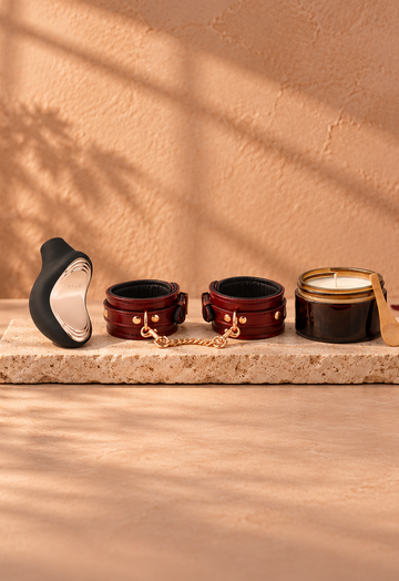 Black LELO pleasure toy, wine-red leather cuffs and massage candle on light travertine, with warm botanical shadows and burgundy fabric