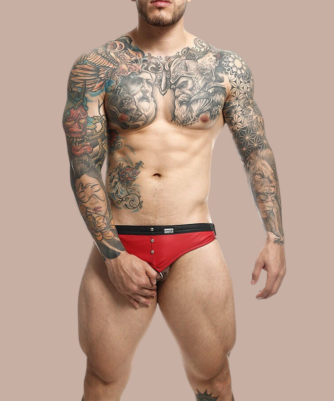 Pleasurements Mob Dngeon jockstrap in rood leer en metaal gedragen door een getatoeëerde man voor een witte achtergrond