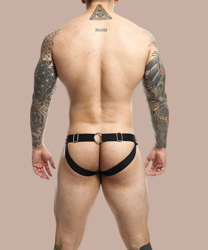Pleasurements Mob Dngeon jockstrap in zwart leer en metaal, gedragen door een getatoeëerde man van achteren gezien.