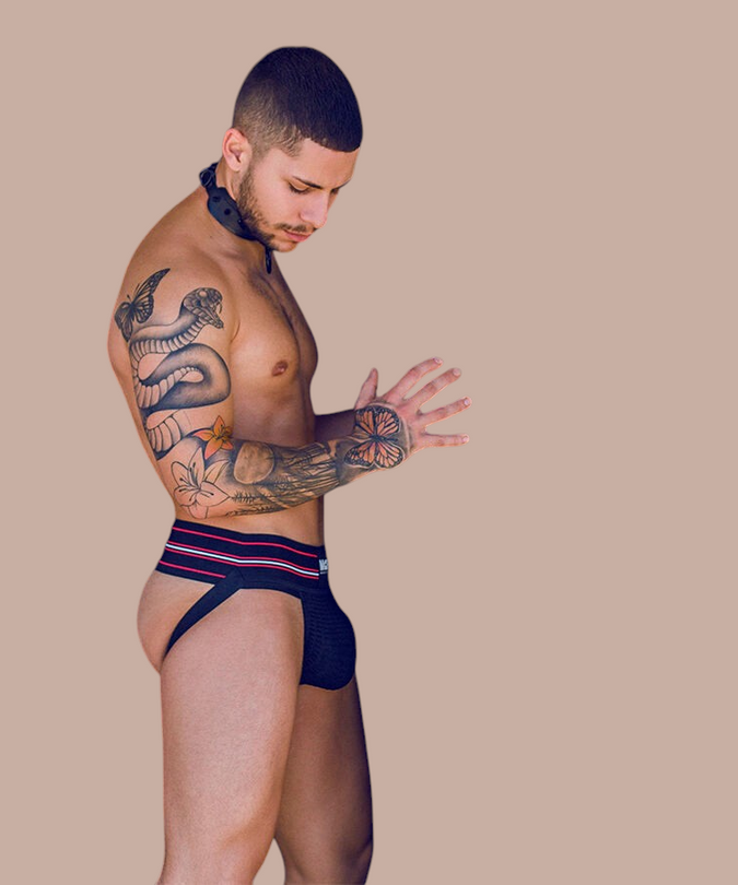 Gemanierde man met tatoeages draagt een zwarte Pleasurements Mob Classic thong met rode en witte accenten