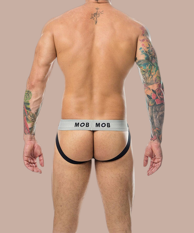 Achterkant van een witte Mob jockstrap met grijze tailleband en zwart logo, gedragen door een getatoeëerde man.