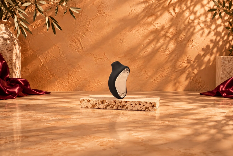 Black LELO SONA 2 on light travertine against a warm copper-beige background with burgundy satin accents