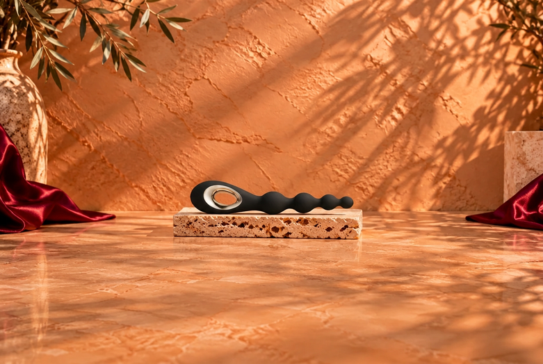 Black anal toy with a metallic ring detail resting horizontally on a light travertine slab, against a warm terracotta background with botanical shadows and burgundy satin