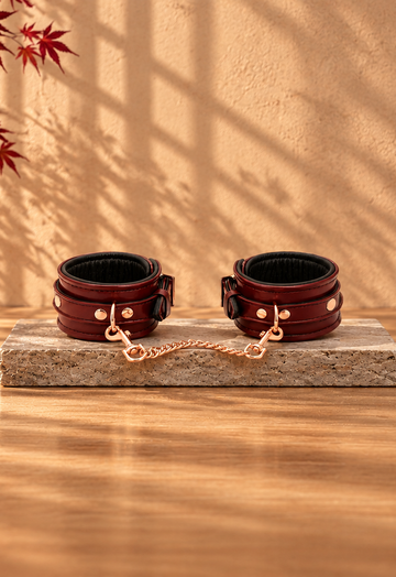 Wine-red leather ankle cuffs with a gold-coloured chain on light stone, with Japanese lattice shadows, red maple leaves and burgundy fabric