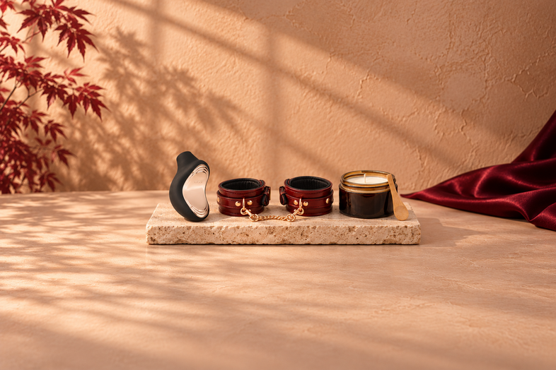 Black LELO pleasure toy, wine-red leather cuffs and massage candle on light travertine, with warm botanical shadows and burgundy fabric