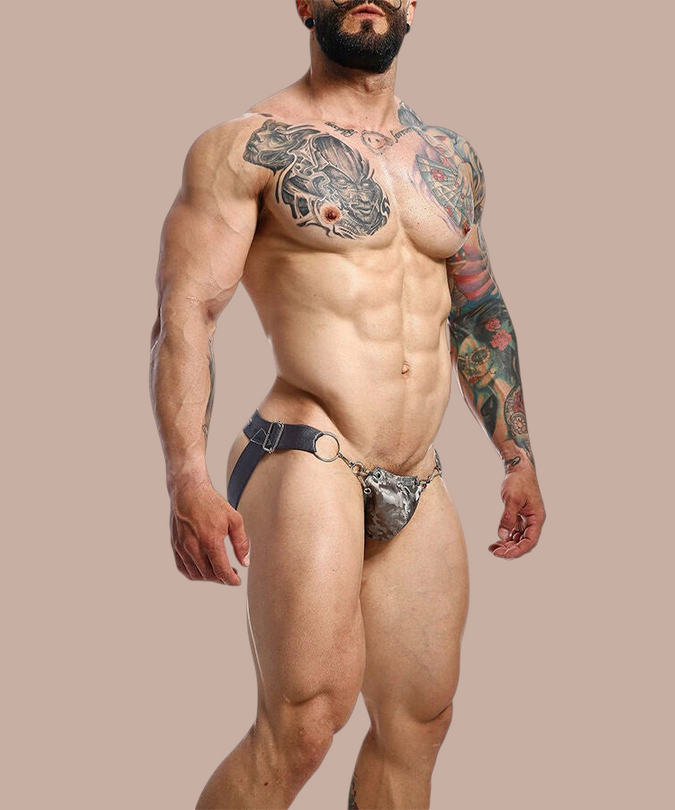 Pleasurements Mob Dngeon Snap jockstrap in grijs leer en kant met metalen ringen gedragen door een getatoeëerde man.
