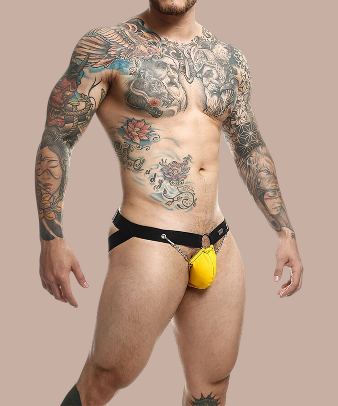 Pleasurements Mob Dngeon gele jockstrap met metalen kettingen gedragen door een getatoeëerde man
