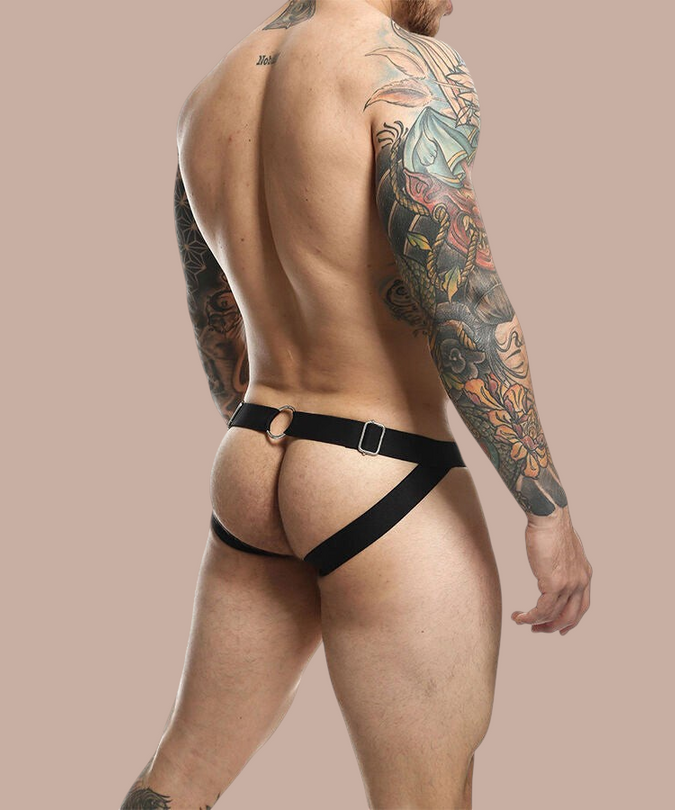 Pleasurements Mob Dngeon zwarte jockstrap met metalen ringen gedragen door een getatoeëerde man op een beige achtergrond