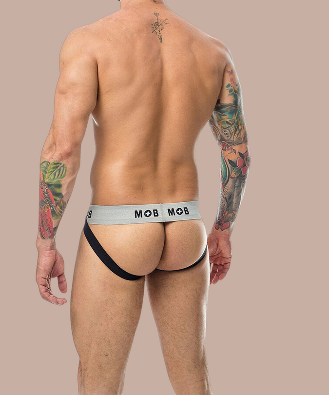 Achterkant van een witte Mob jockstrap met elastische tailleband van Pleasurements gedragen door een getatoeëerde man