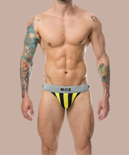 Pleasurements Mob Classic Jock Stripe in geel met grijze tailleband gedragen door een man met getatoeëerde armen